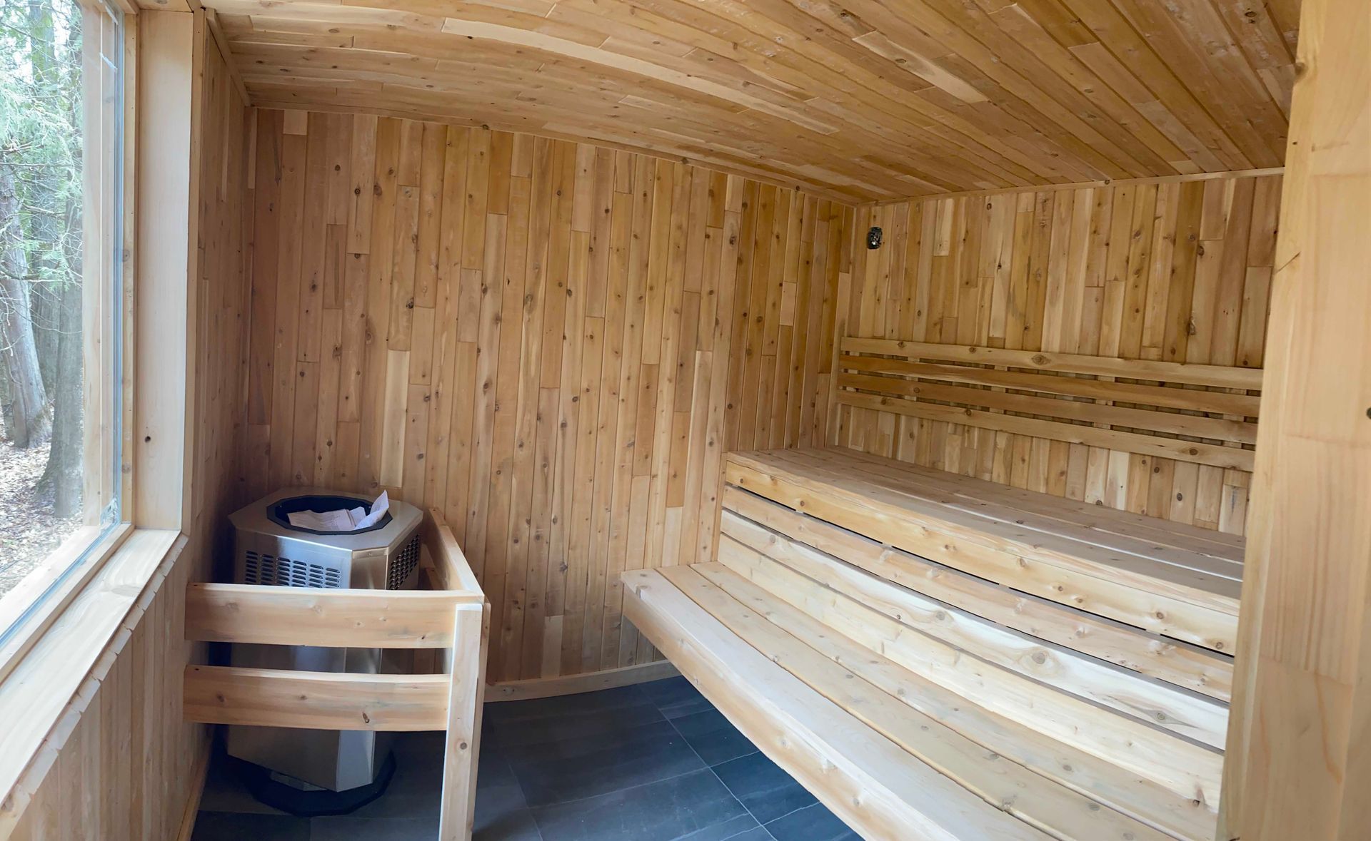 Un sauna intérieur avec des murs lambrissés, des bancs en bois à plusieurs niveaux, un poêle en métal rempli de pierres et une grande fenêtre latérale.