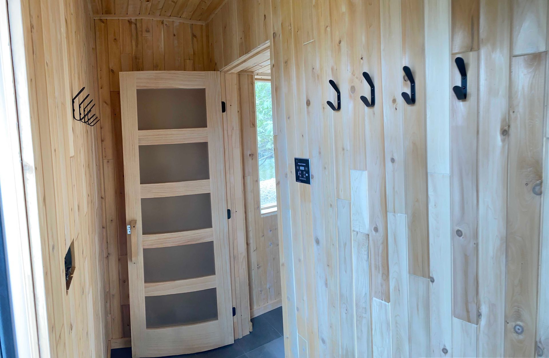 Un couloir en bois clair avec des lambris verticaux, des patères noires fixées au mur et une porte à cinq panneaux dépolie.