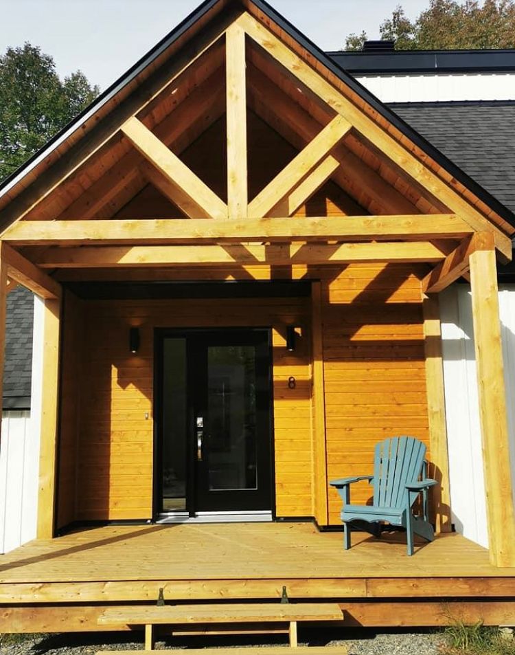 Un porche d'entrée en bois avec un toit à pignon, une porte d'entrée noire et une simple chaise Adirondack bleu clair sur la terrasse.