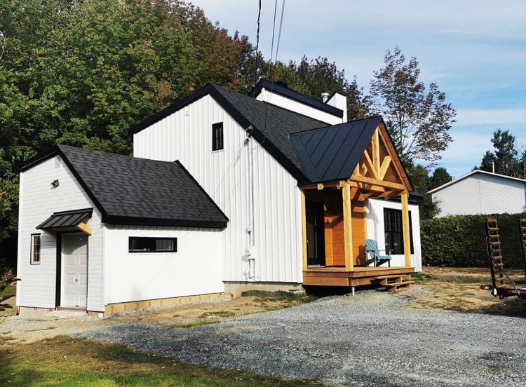 Une maison blanche moderne avec des finitions et un toit noirs, un bardage vertical, un porche à ossature bois et une allée en gravier.