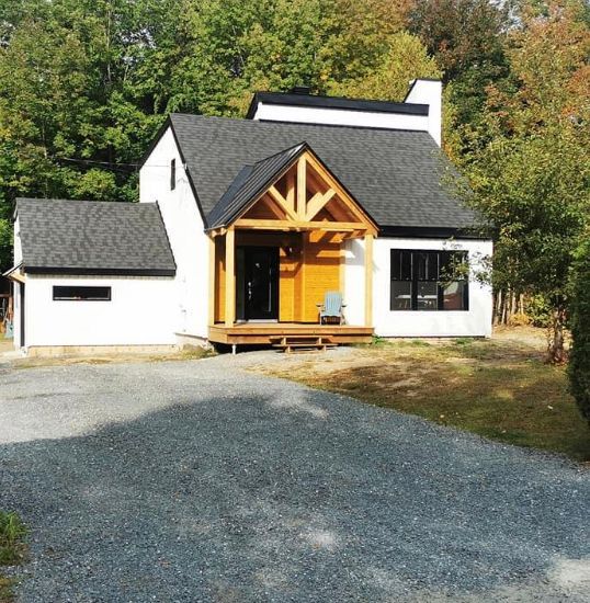 Une maison blanche au toit noir, avec un porche à ossature bois et une allée de gravier, entourée d'arbres par une journée ensoleillée.