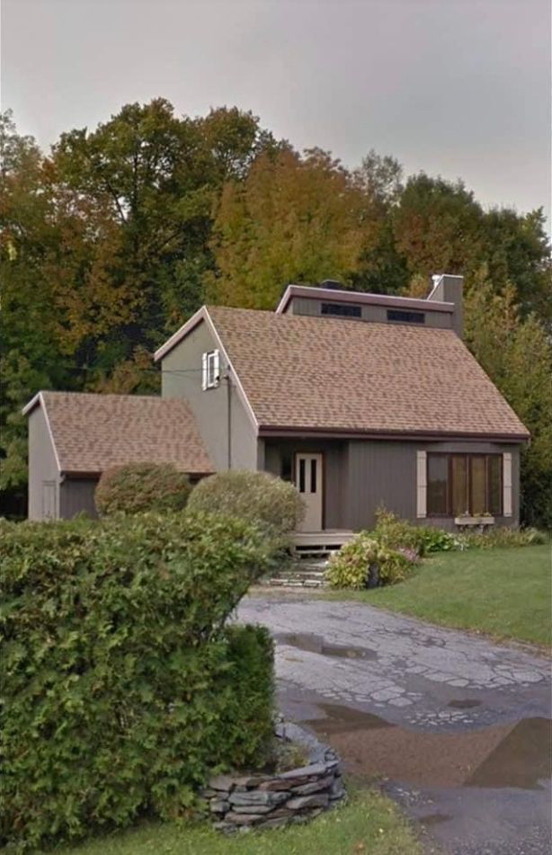Une maison grise à deux étages, avec un toit en bardeaux bruns, nichée derrière une haie verte et des arbres, sur une allée pavée.