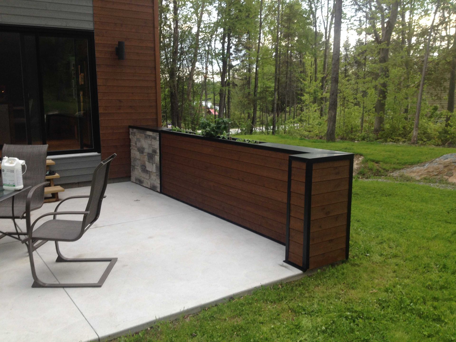 Un muret en bois orné de pierres préserve l'intimité d'une maison, devant laquelle se dresse un patio en béton, à côté d'un jardin boisé.