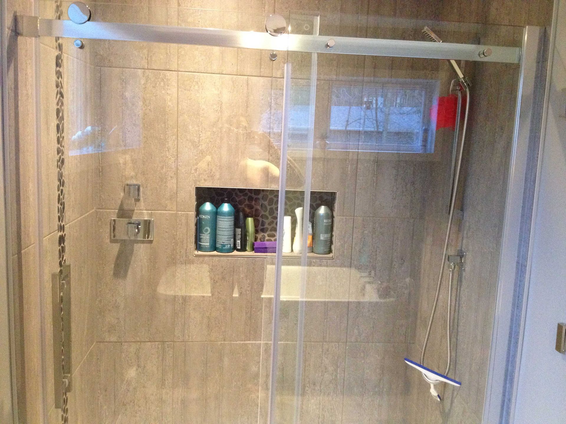 Une douche à l'italienne vitrée avec des murs carrelés beiges, une niche de rangement centrale et un mécanisme de porte coulissante.