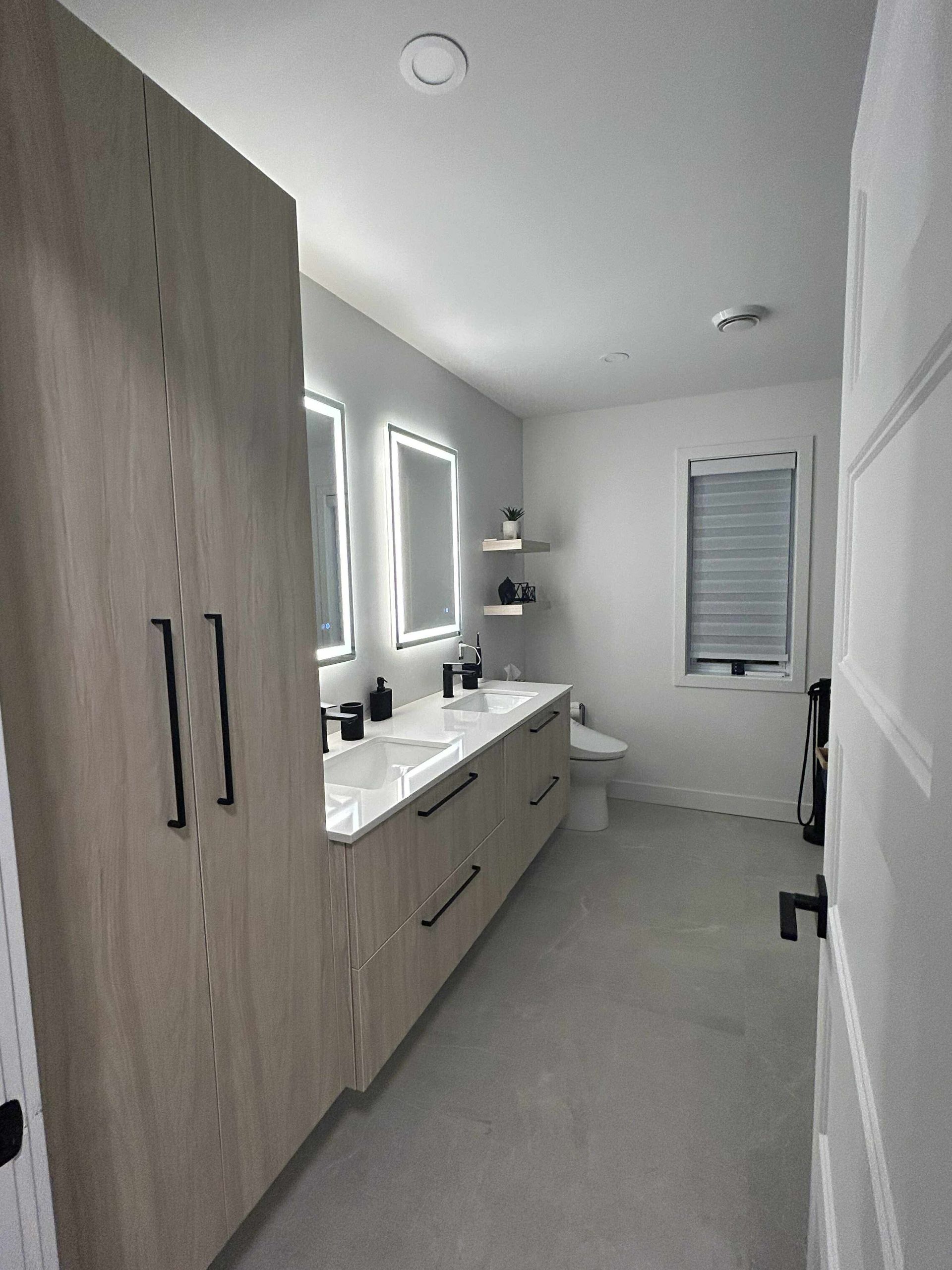 Une salle de bains moderne avec un meuble-lavabo en bois clair, deux miroirs éclairés, des étagères flottantes et un sol carrelé gris.