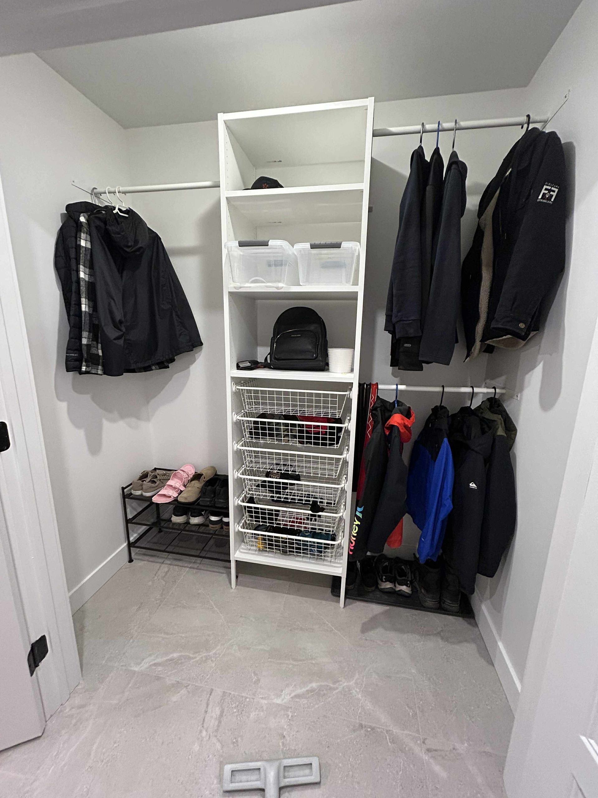 Un dressing avec des portants pour les manteaux, une étagère centrale avec des bacs de rangement et un range-chaussures au sol.