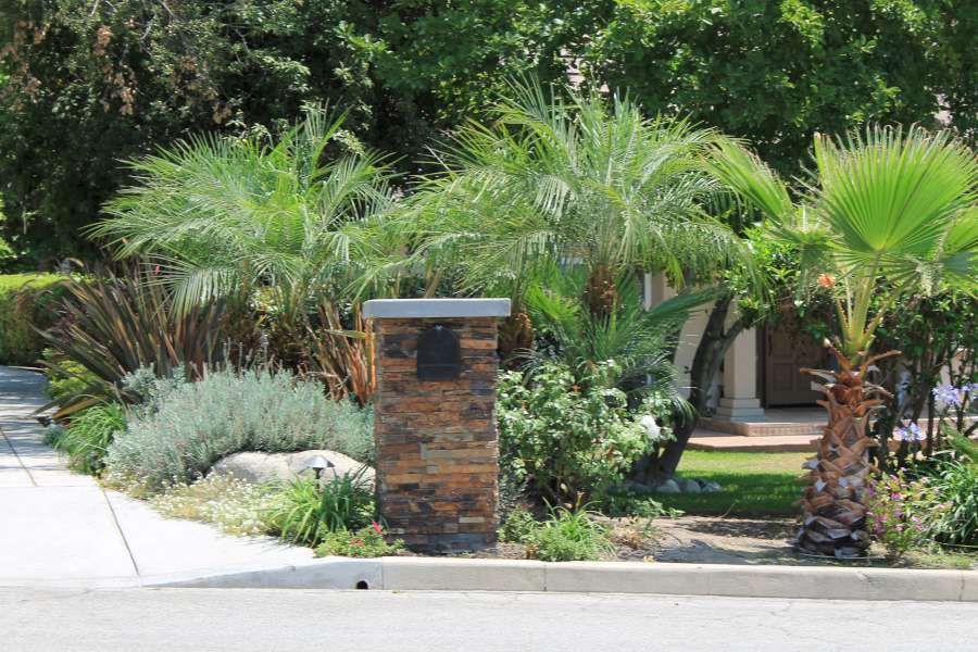 A brick pillar with a mailbox in front of a house surrounded by palm trees.