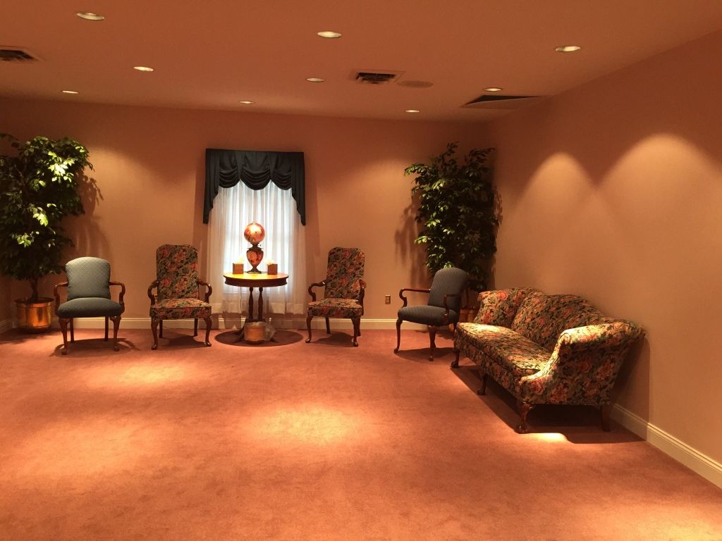 A sparsely furnished waiting room with chairs and a sofa, likely in a funeral home. Warm lighting and a window with a decorative valance.