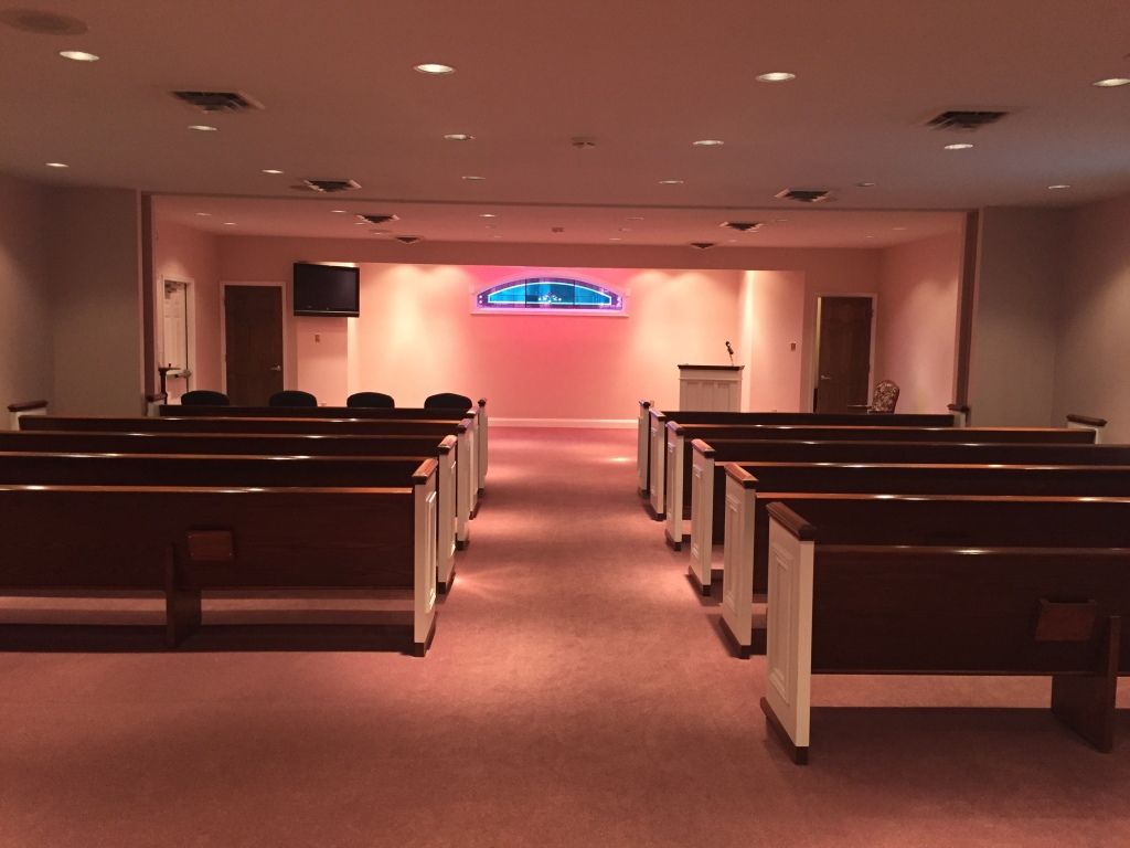 An empty chapel with rows of wooden pews facing a raised platform. The space has pink carpeting and lighting.