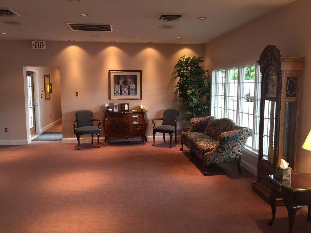 Interior of a funeral home waiting area with a couch, chairs, and a grandfather clock. Pink carpet and walls, a window, and a tree are also visible.
