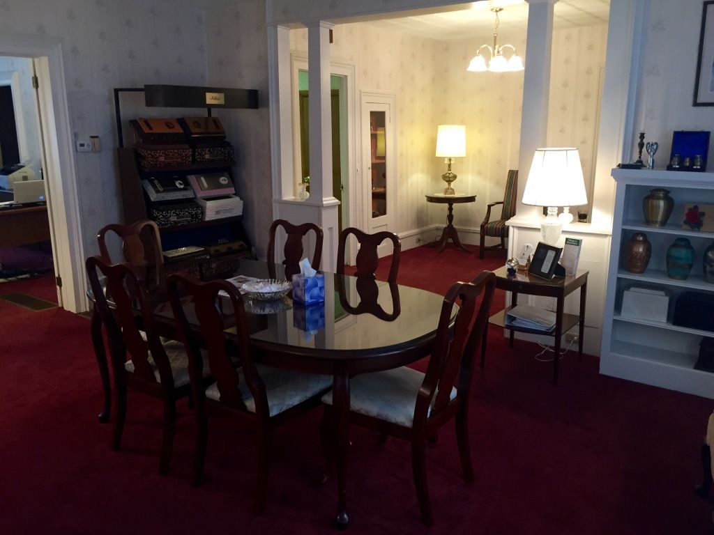 A room with a dining table and chairs, red carpet, and a view into a room with a lamp and seating. A display shelf is visible.