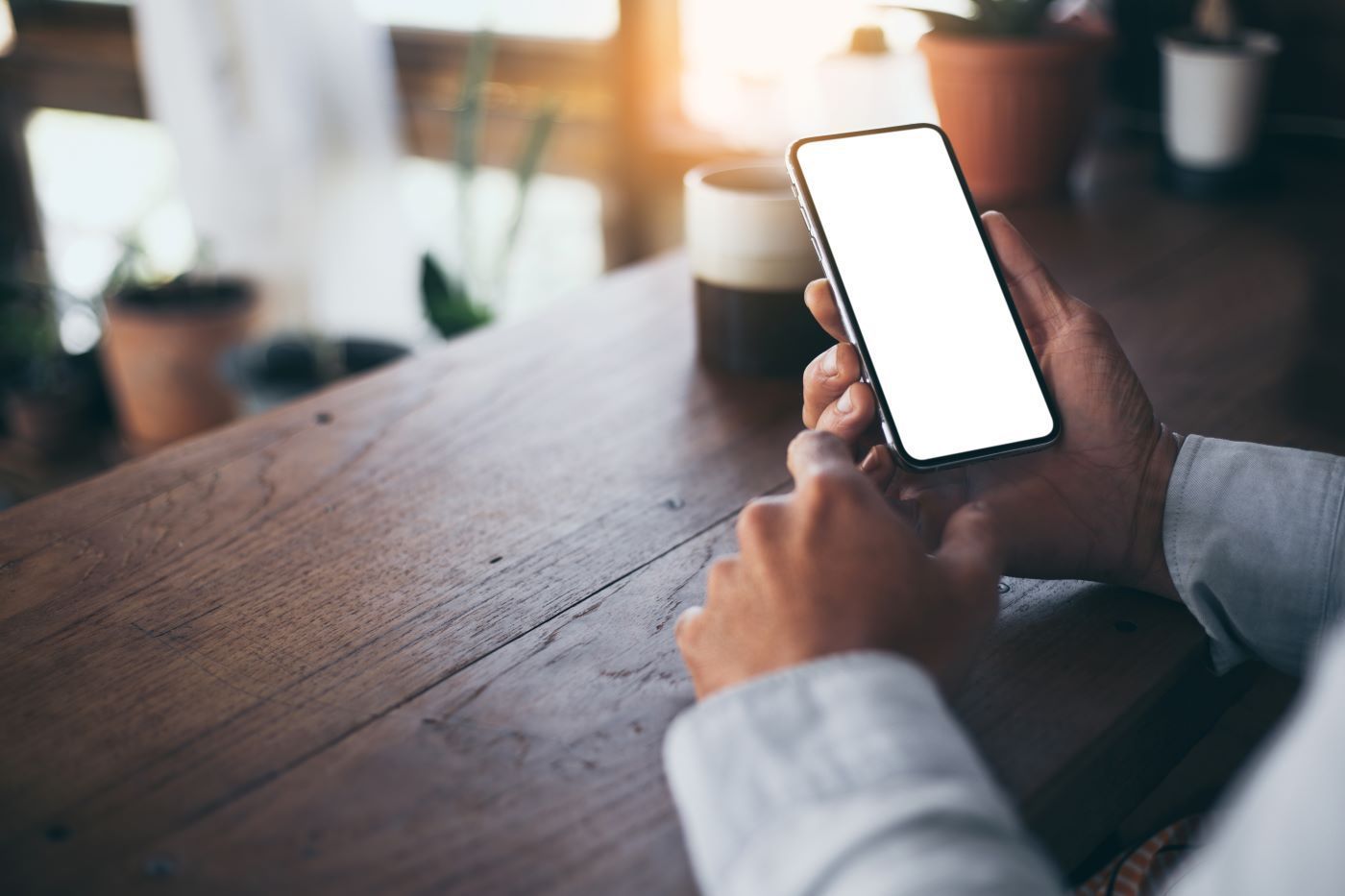 Person holding a smartphone with a blank white screen, sitting at a wooden table near a window with plants and a mug.