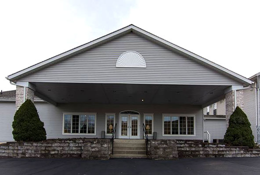 Gray building with a covered entrance, double doors, and windows. Two green bushes flank the entrance.