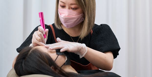 A woman wearing a mask and gloves is applying makeup to a woman 's eyebrows.