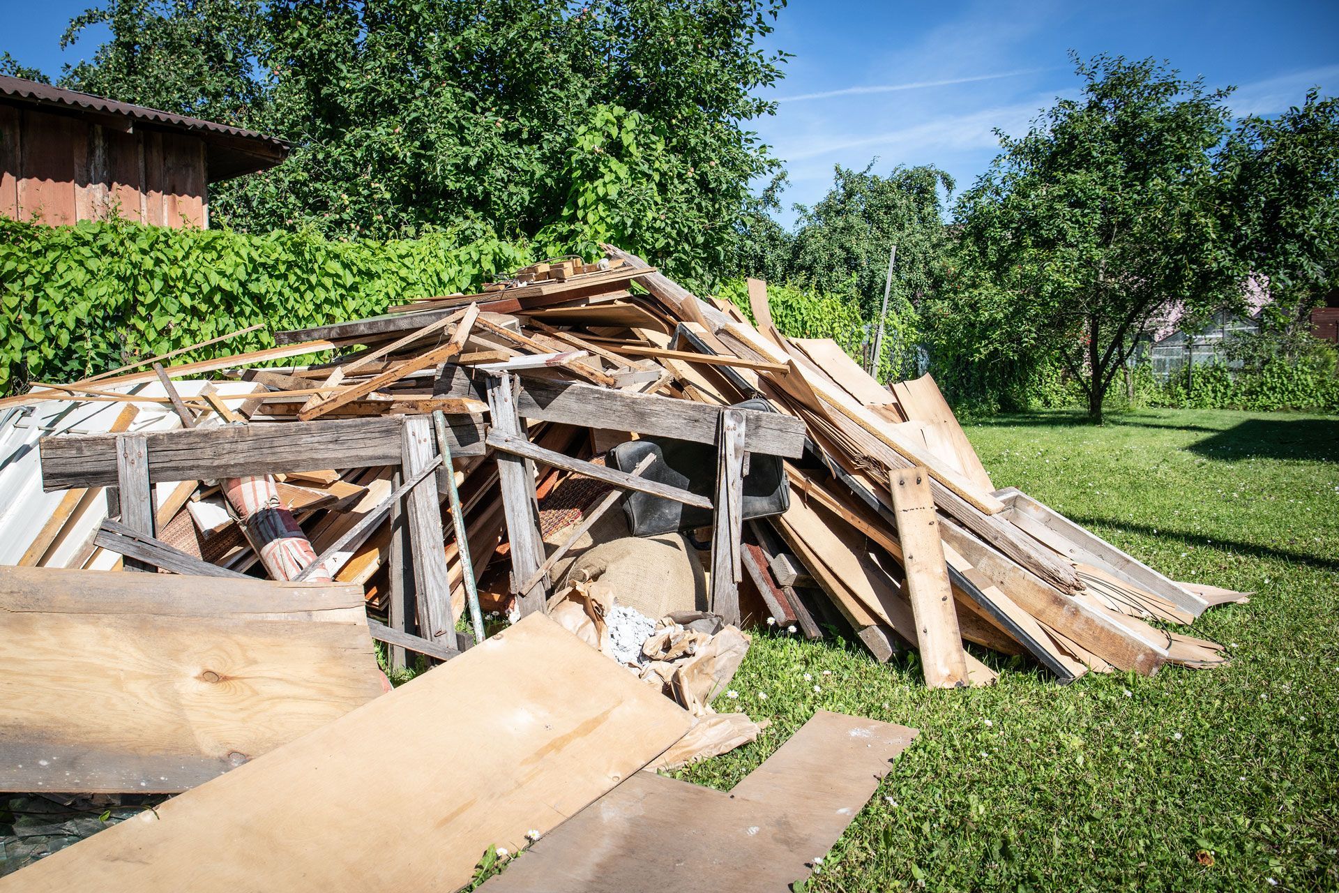 Pile of broken wooden debris in a grassy yard with a house and trees in the background