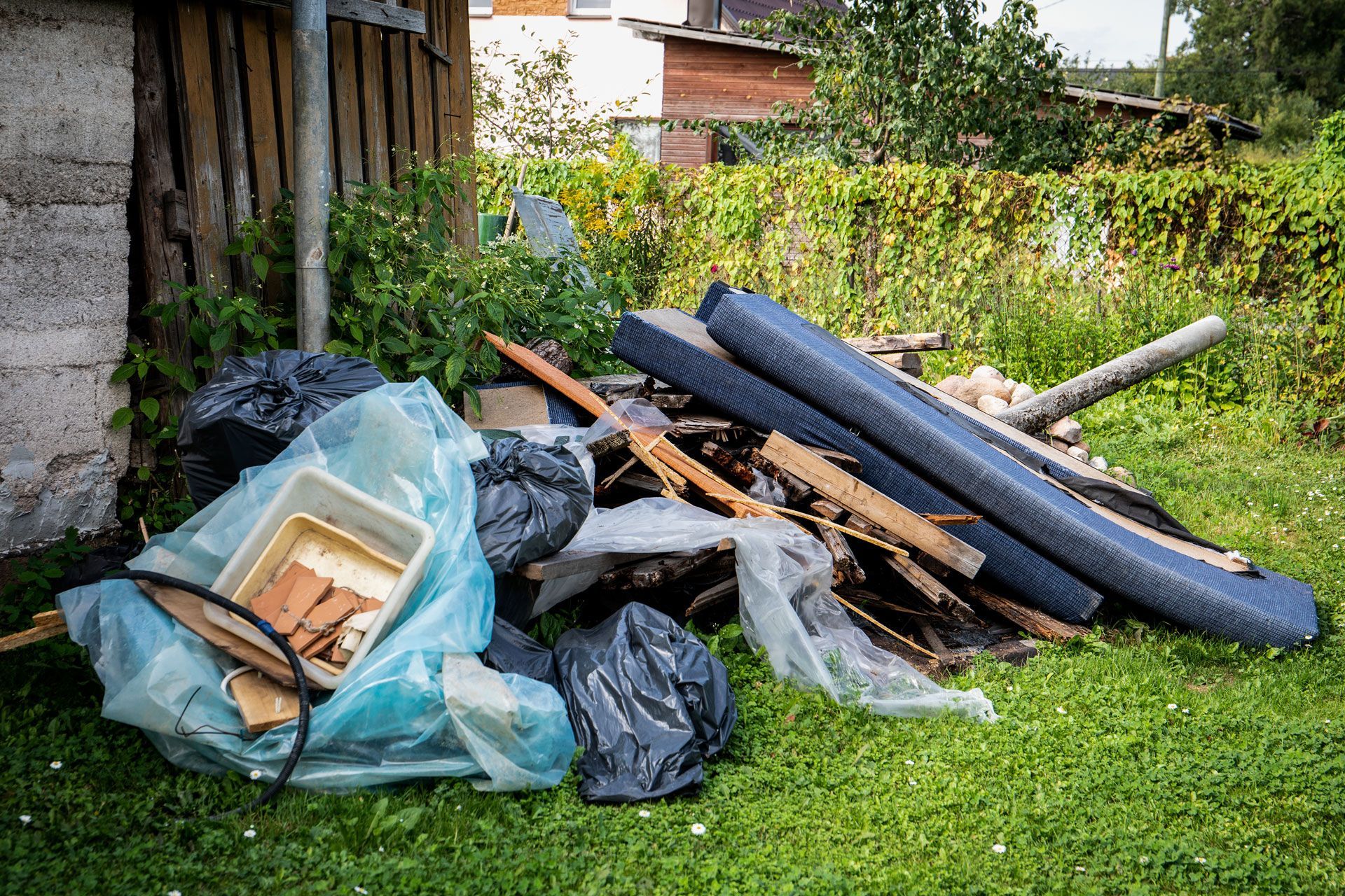 Pile of discarded furniture, tires, and trash bags on grass beside a shed in a yard