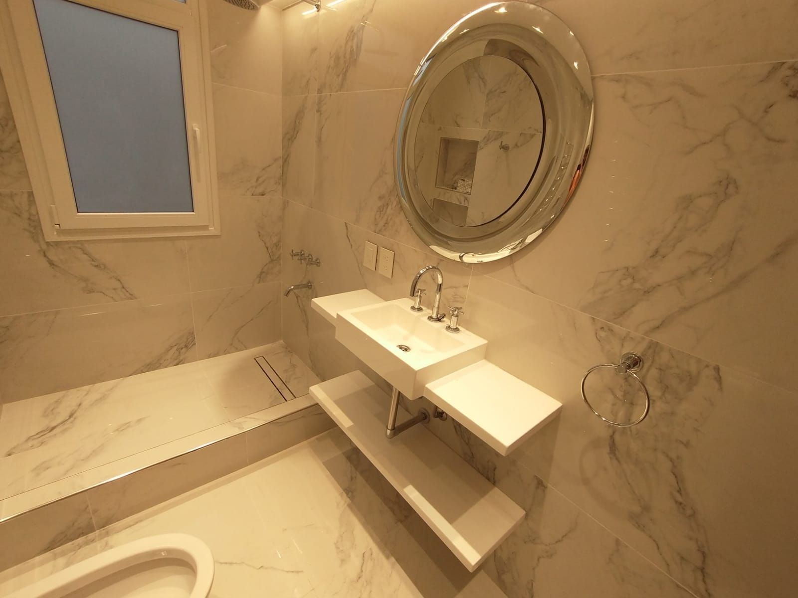Baño moderno con paredes de mármol blanco, espejo redondo y lavabo flotante.