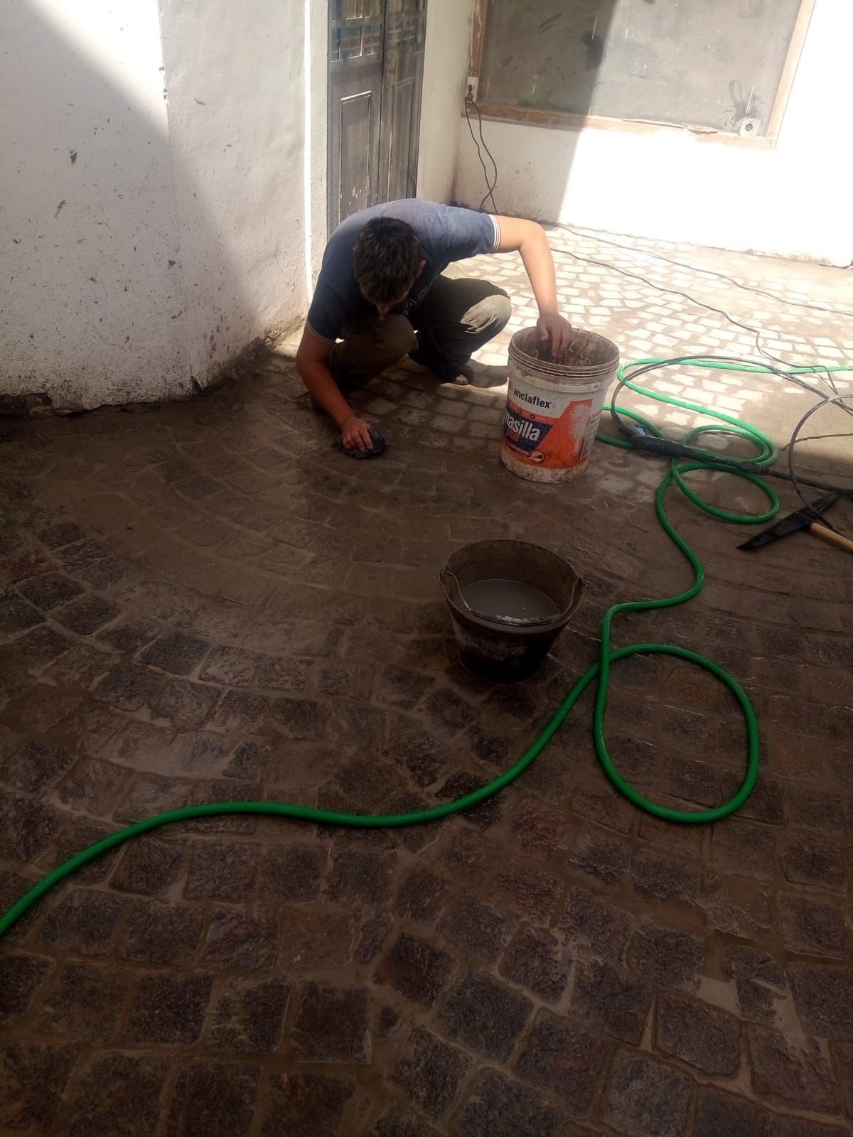Hombre aplicando lechada a un patio adoquinado, usando un balde y una manguera.