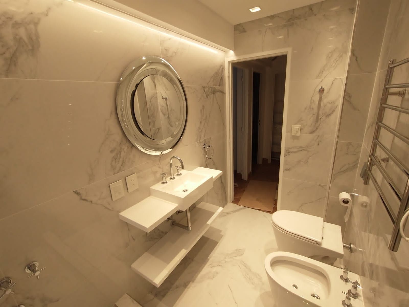 Baño moderno con paredes de mármol blanco, espejo redondo y sanitario.