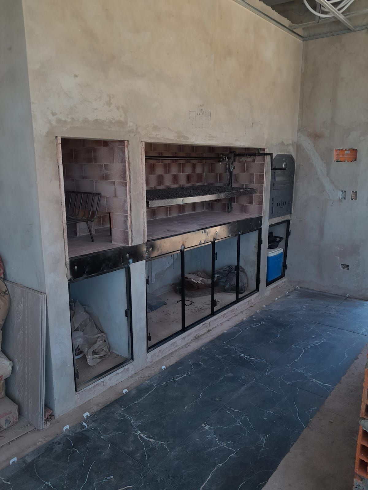 Una cocina al aire libre parcialmente construida con horno de ladrillo, detalles de metal y piso de concreto.