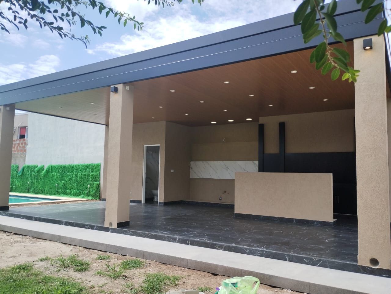 Patio cubierto al aire libre, paredes beige y techo de madera; al fondo se ven zonas verdes y una piscina.