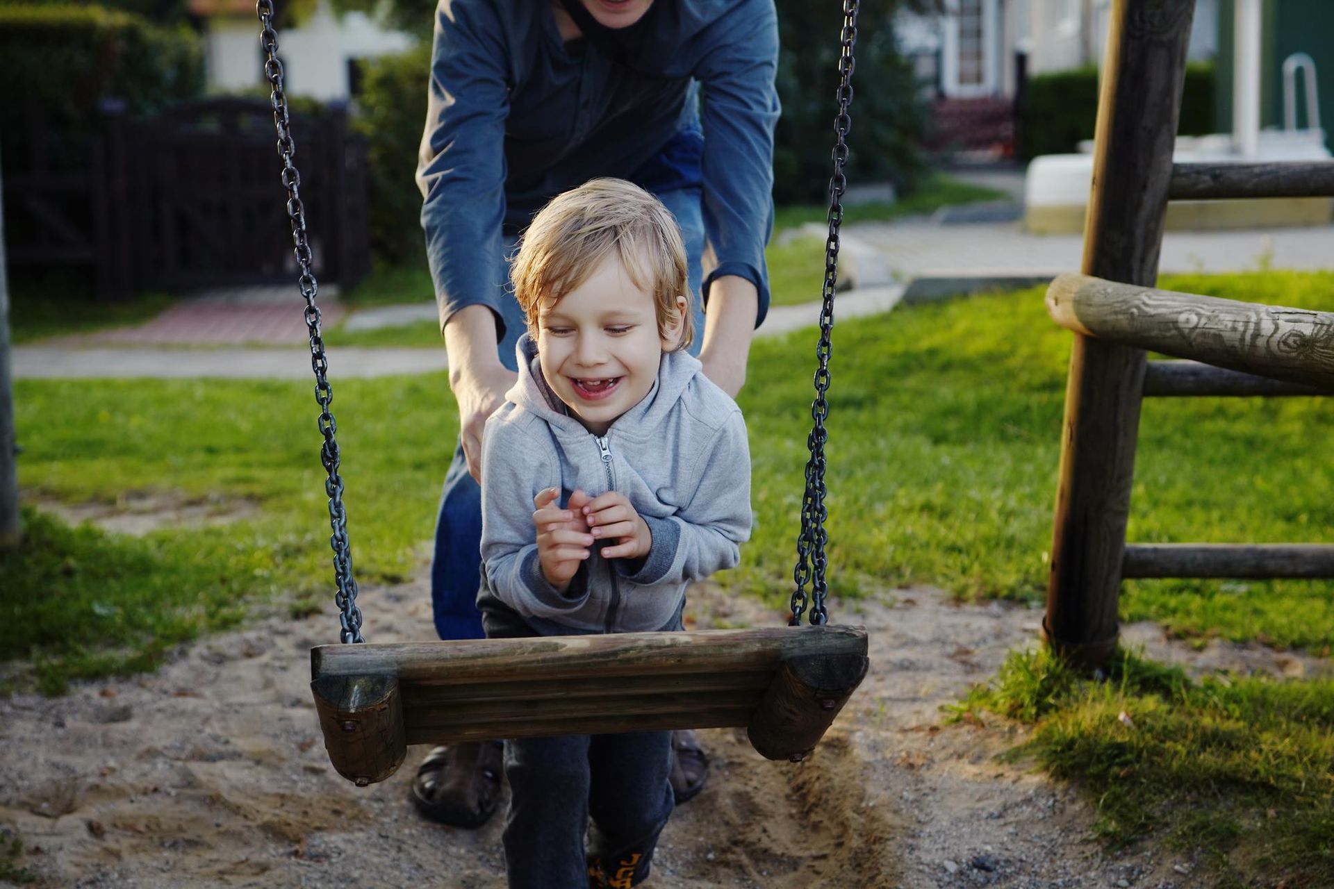 Boy on swing, smiling, being pushed by a person in a park.