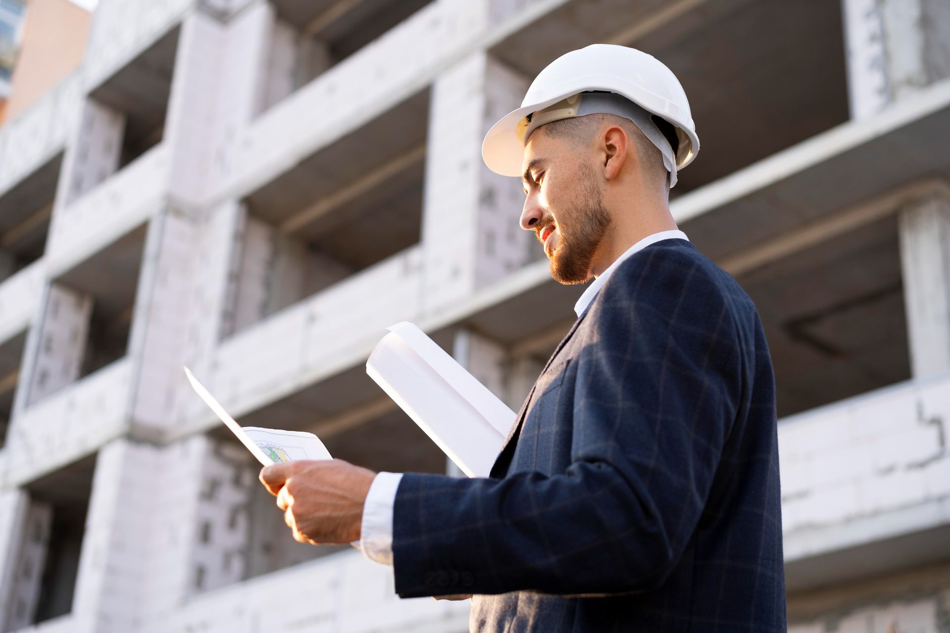 Construction worker in a hard hat and suit examines blueprints at a building site.