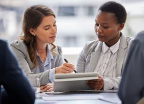 Two women in business attire reviewing documents, discussing details in an office setting.