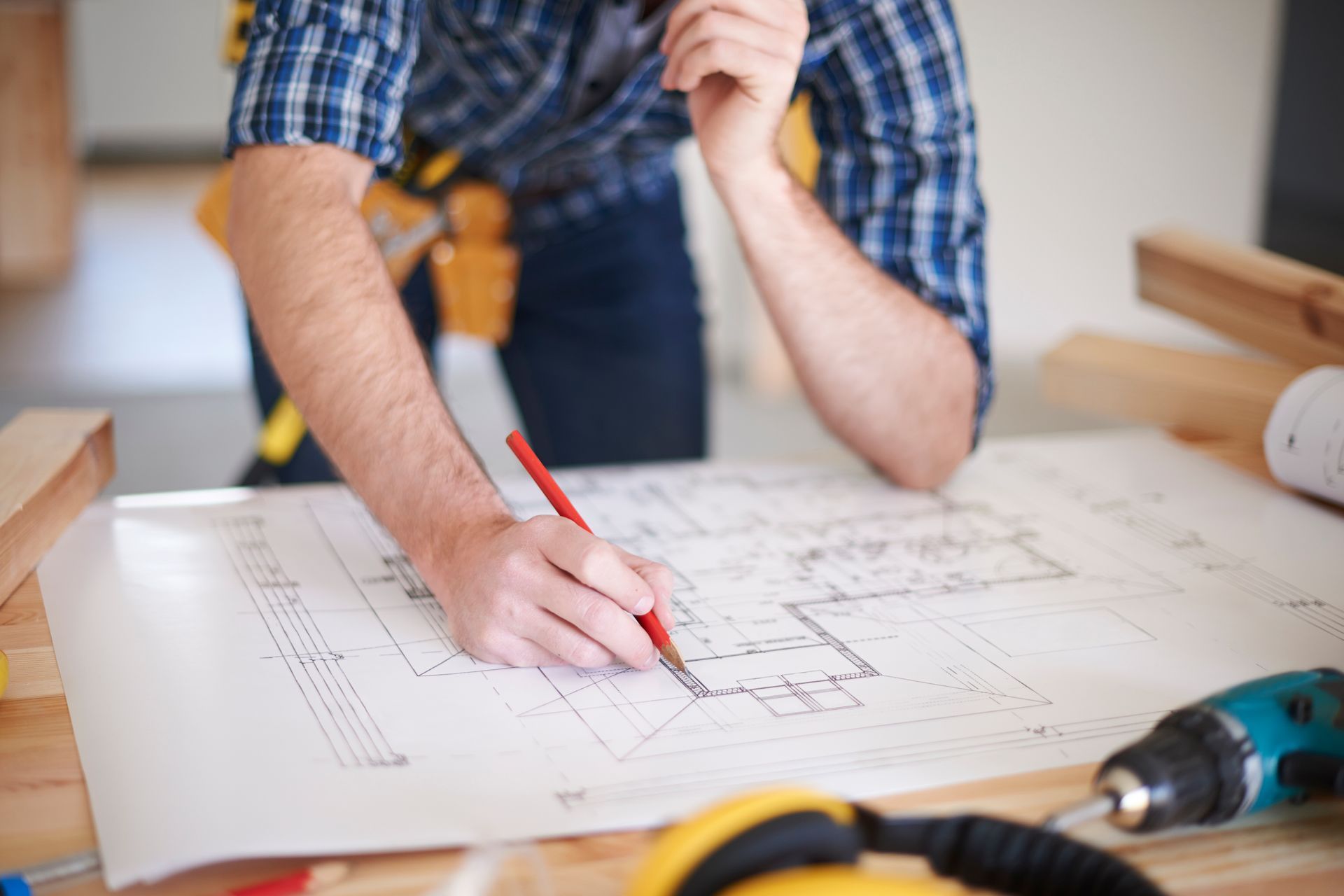 A person in a blue plaid shirt sketches on architectural plans with a red pencil at a wooden table.