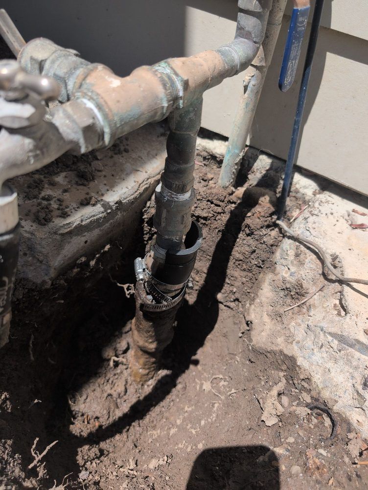 Excavated outdoor water pipe with a break. Bronze pipes against a beige siding and dirt.