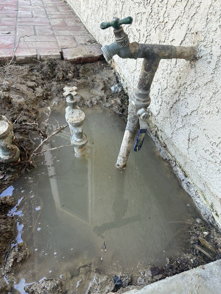 Muddy ground pooled with water around two outdoor faucets on a stucco wall, near brick patio.