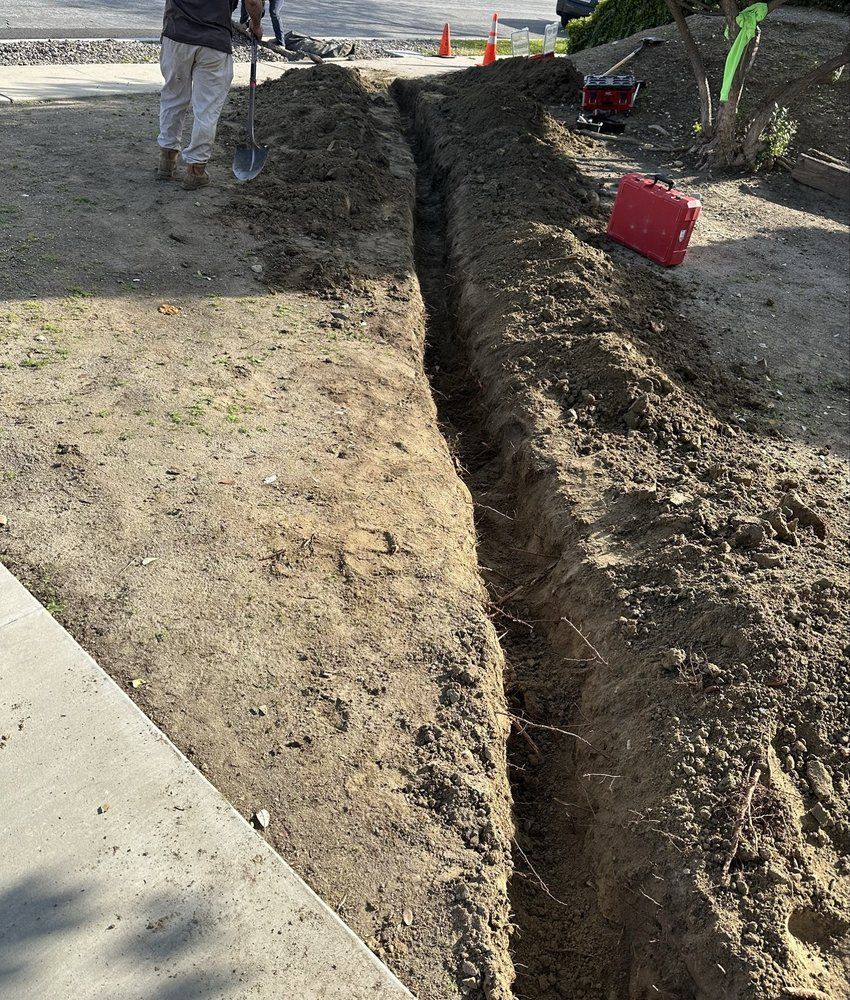 A trench dug in dirt alongside a sidewalk. A worker stands nearby with a shovel.