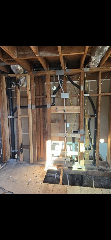 Interior of a home under construction, showing exposed framing, pipes, electrical wiring, and a hole in the floor.