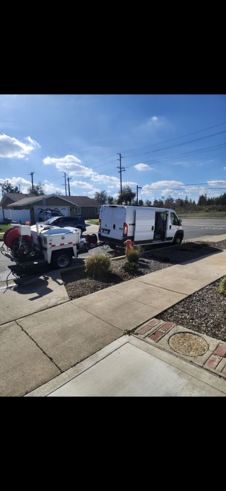 A white van with open back doors and a trailer parked in front of a house on a sunny day.