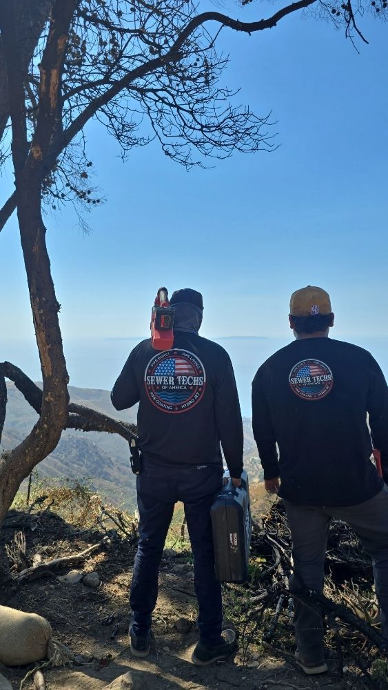 Two men in black shirts with logos stand on a mountain, looking out at a blue sky and landscape.