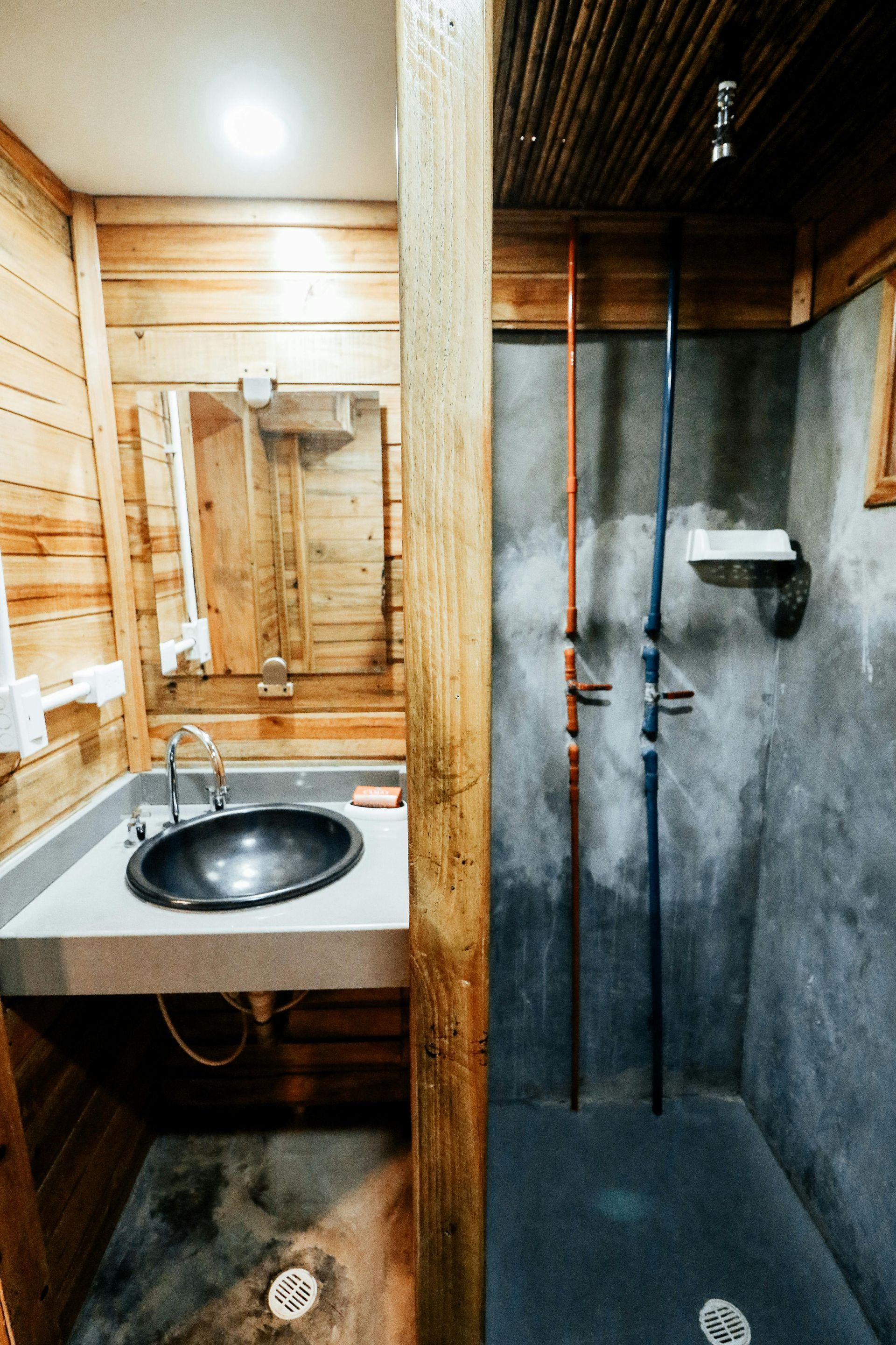 Rustic bathroom: wooden walls, concrete sink, and shower area with exposed pipes.