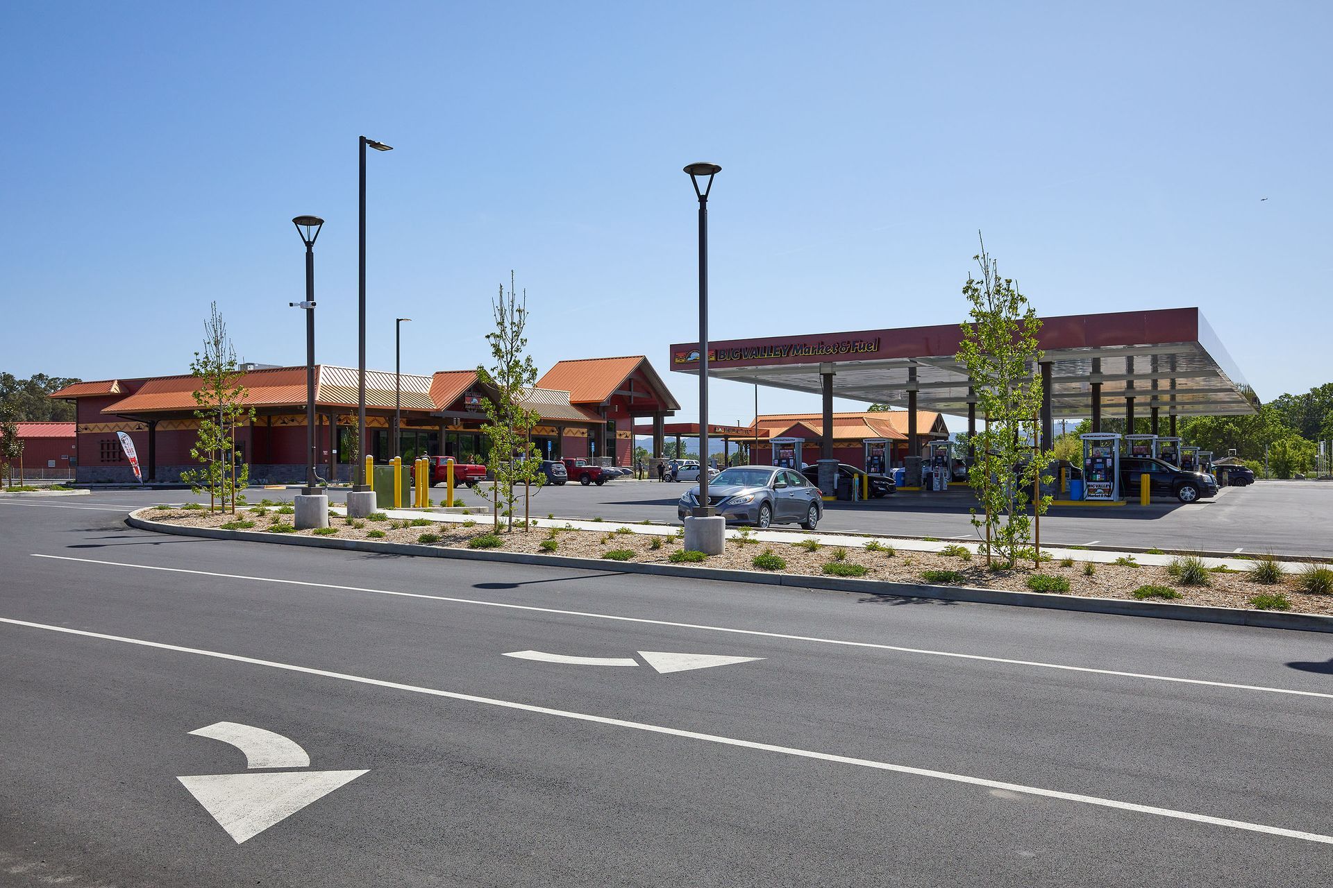 Wide road leading to a modern building complex with parking lot, lampposts, and landscaped median in bright sunlight
