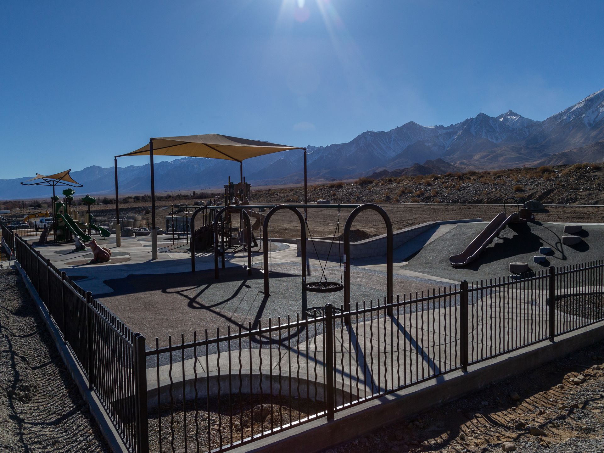Playground with a shade structure, black fence, and mountains in the background on a sunny day.