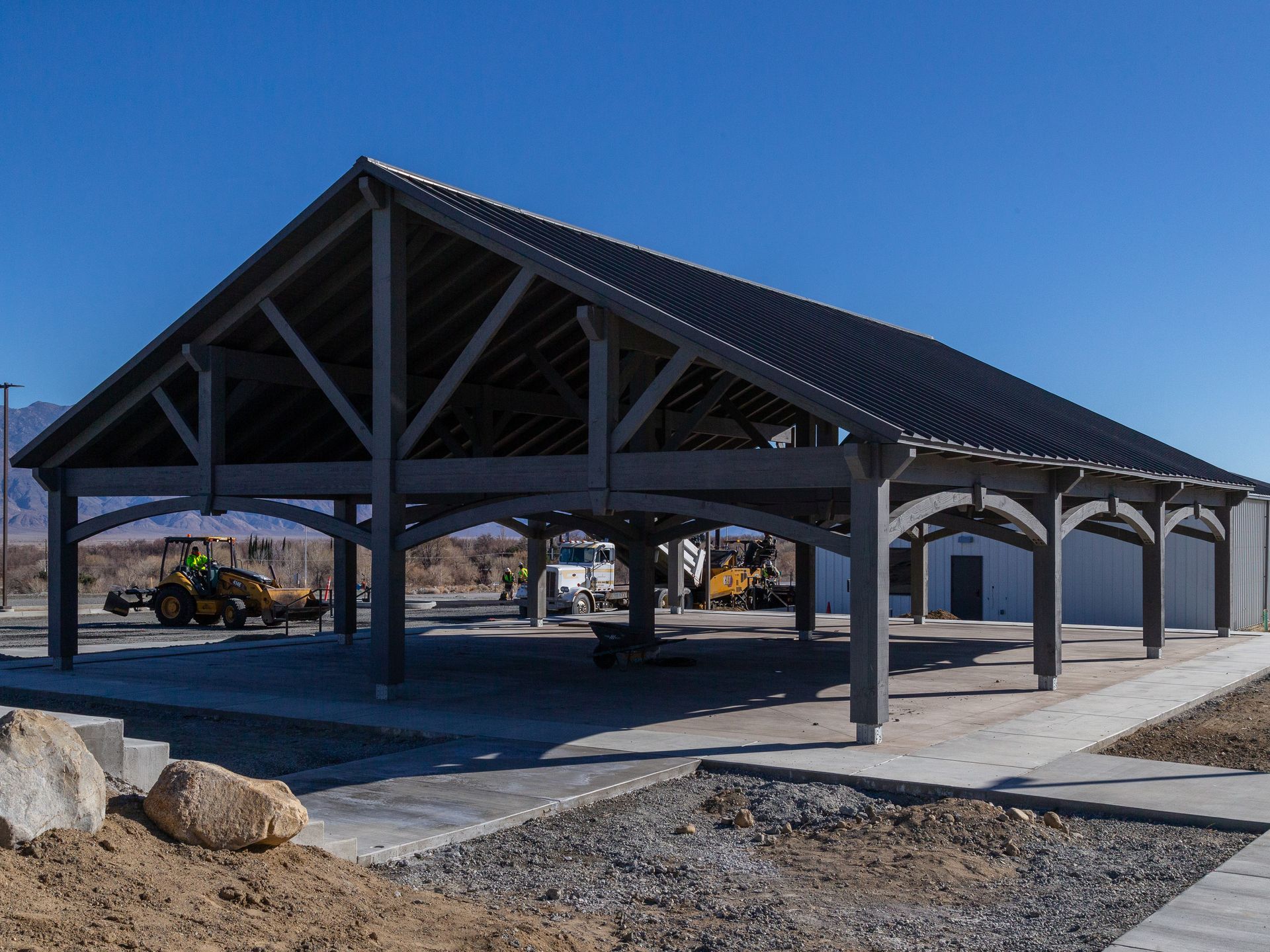 Gray wooden pavilion under construction with a dark roof, parked equipment in the background.
