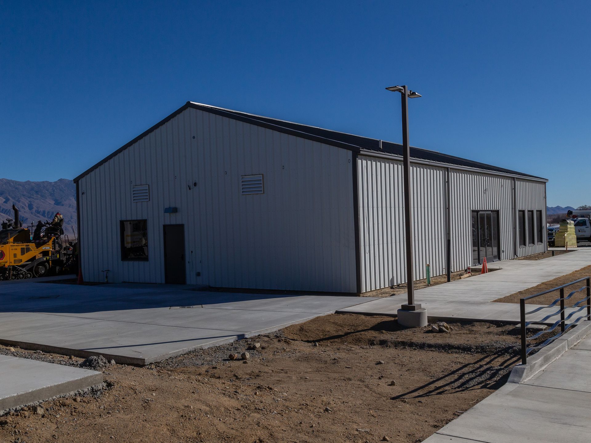 White metal building with dark door and windows, surrounded by a concrete walkway and brown dirt under a clear blue sky.