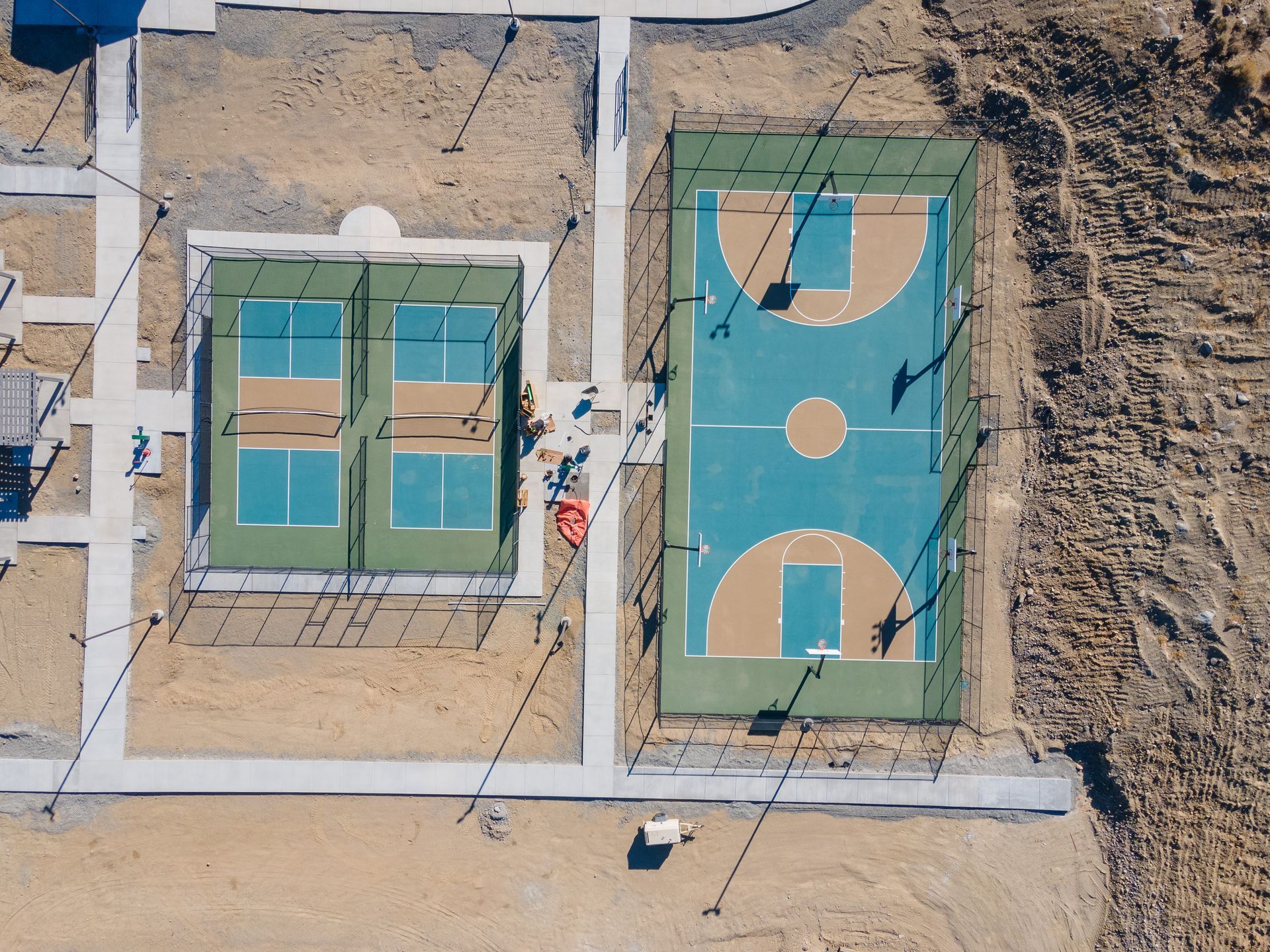 Overhead view of a basketball court and two pickleball courts, all with green and blue surfaces, surrounded by dirt and sidewalks.