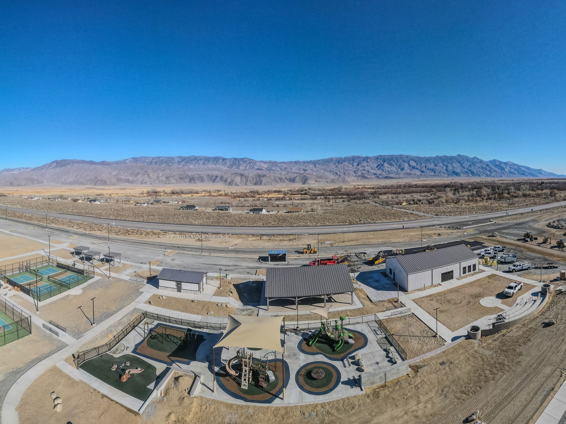 Aerial view of a park with playground, shaded structures, and desert landscape with mountains under a blue sky.