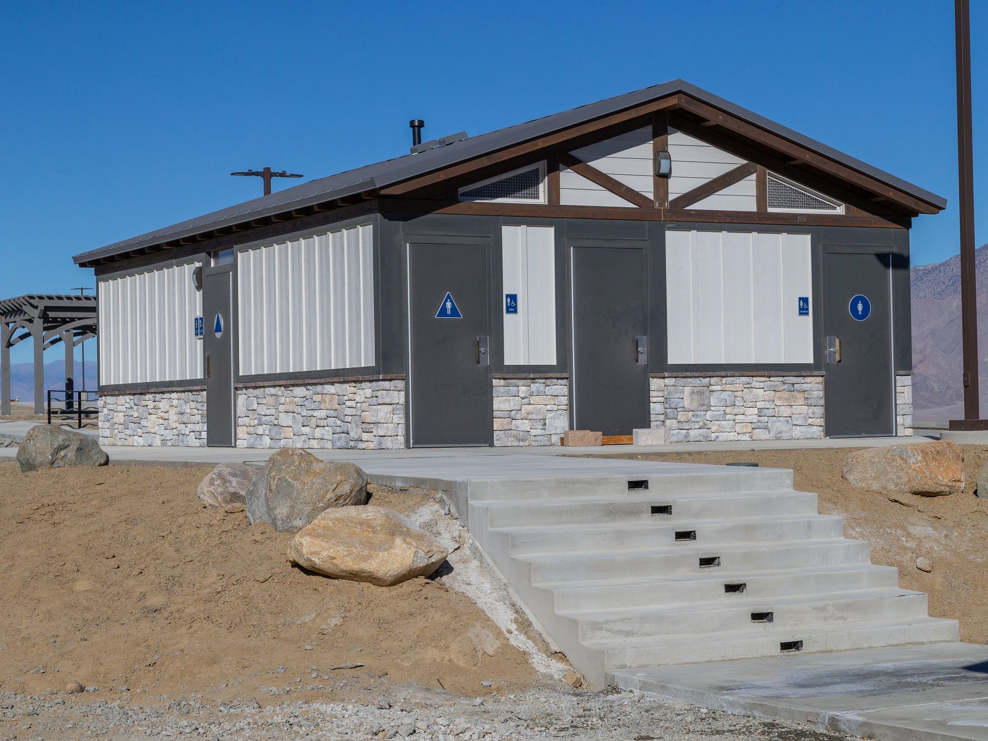 Public restroom with stone base, gray walls, brown roof, blue accessible signs, and concrete steps.