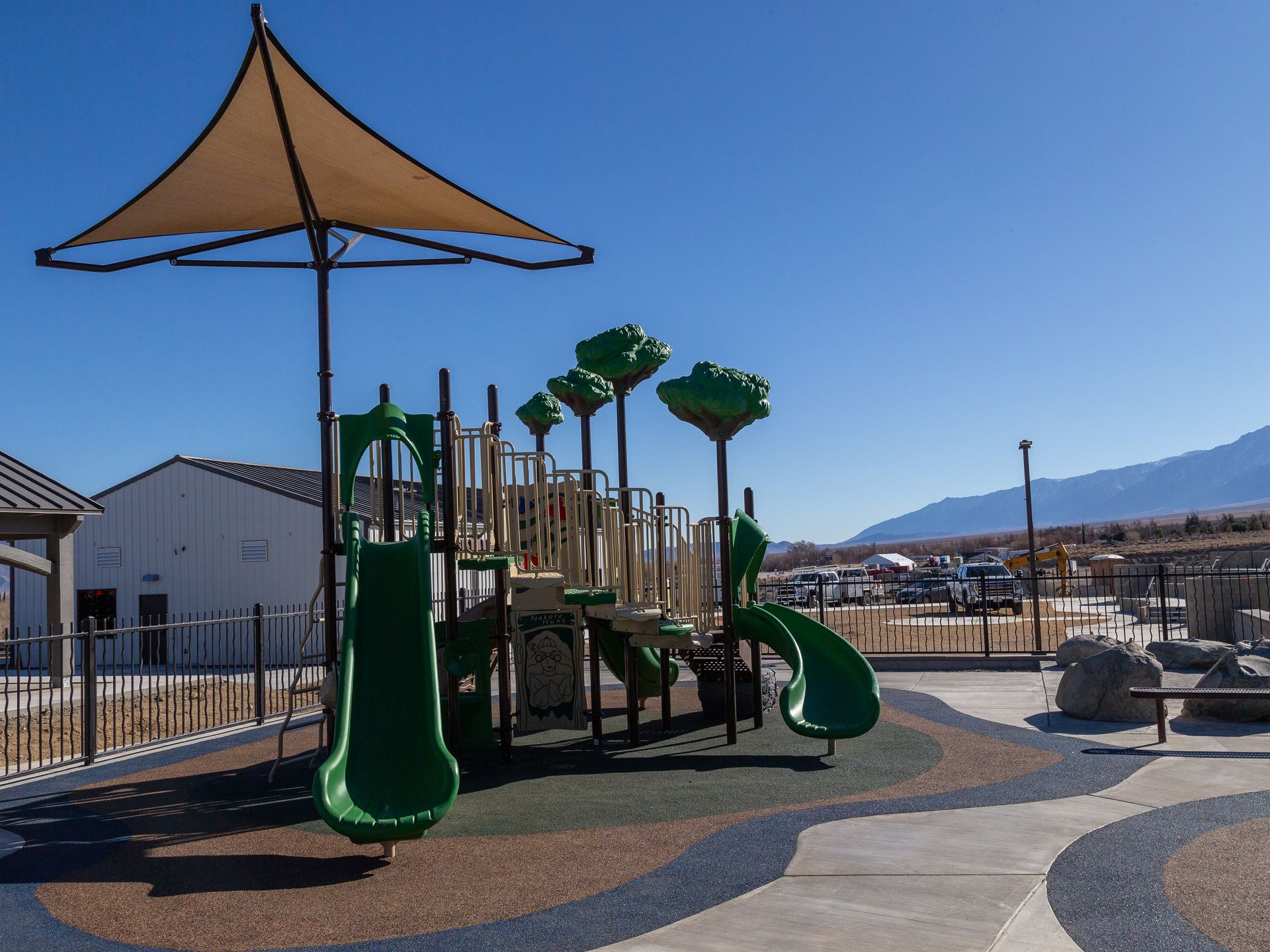 Playground with green slides, shade canopy, and surrounding pathways on a sunny day.