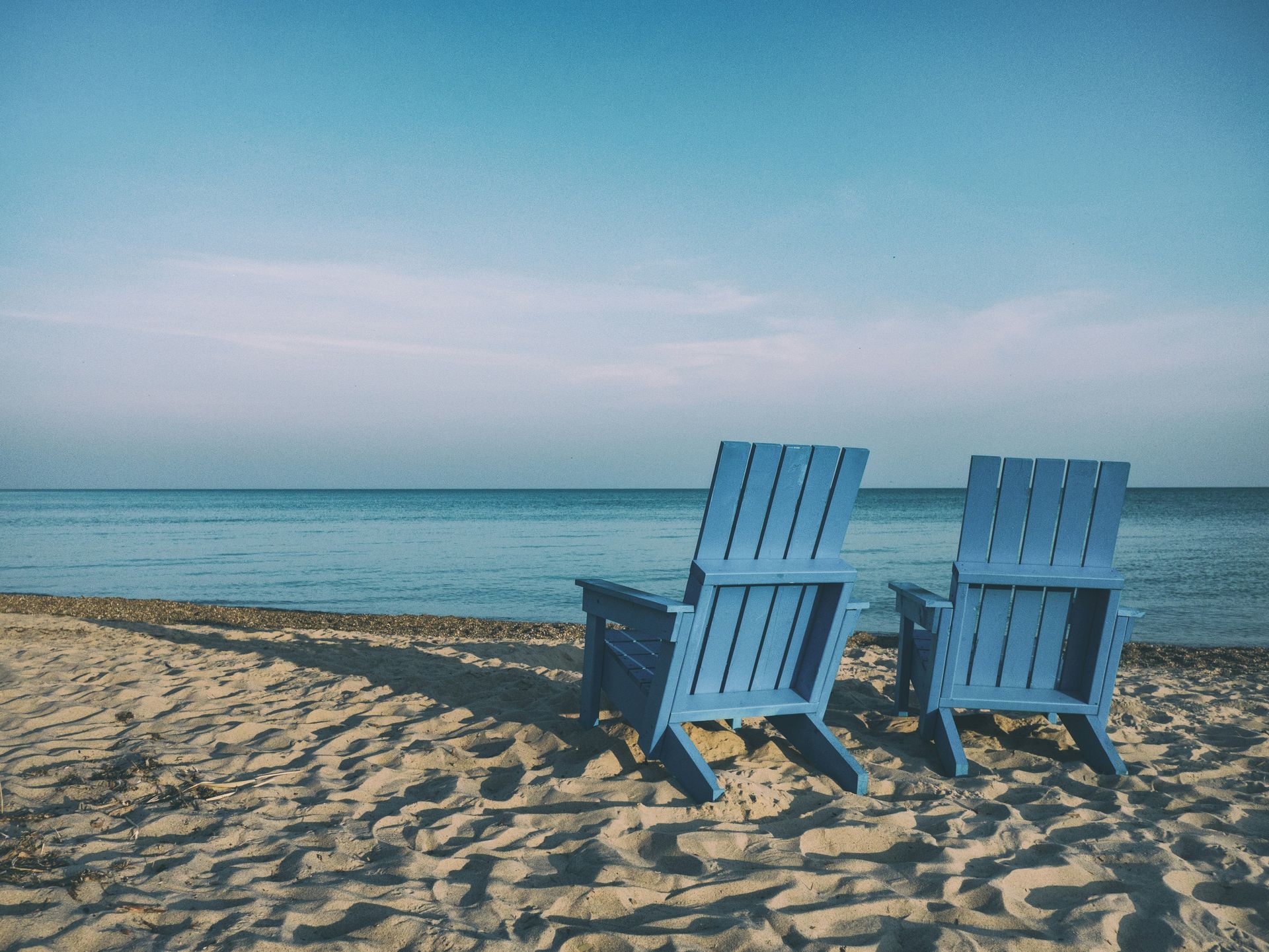 Two blue Adirondack chairs on a sandy beach, facing a calm blue ocean under a clear sky.