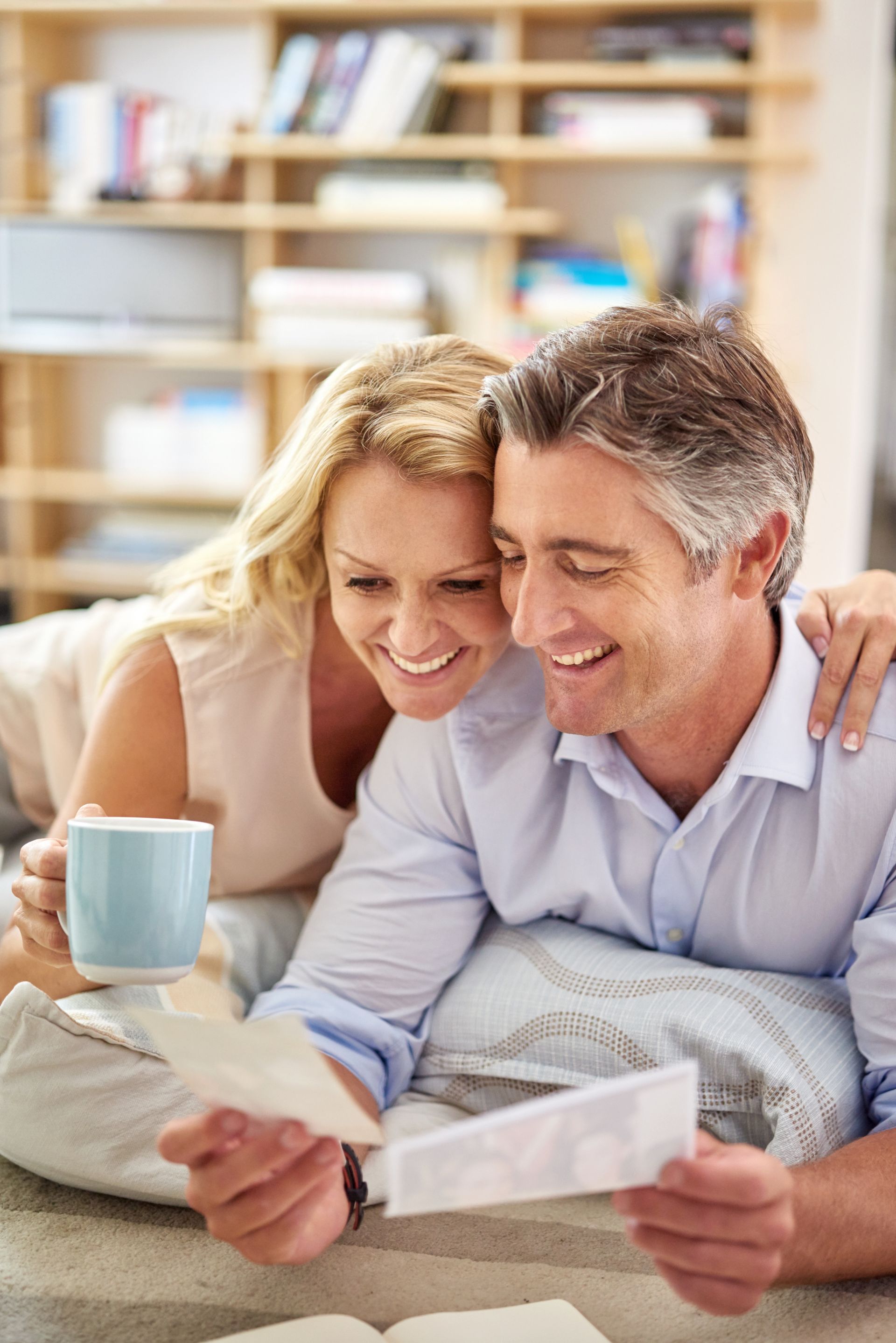Couple smiling, looking at photos together, holding mug, on a bed with a bookshelf in the background.