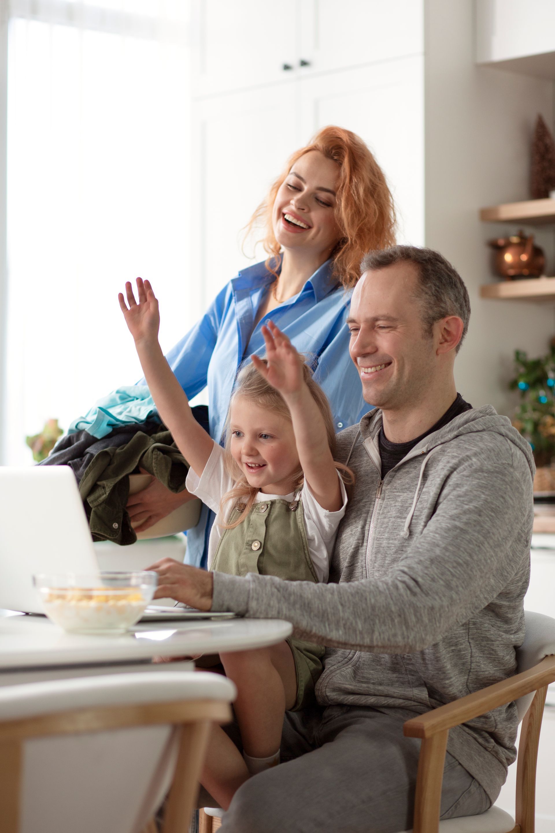 Family of three waves at a laptop screen in a bright kitchen.