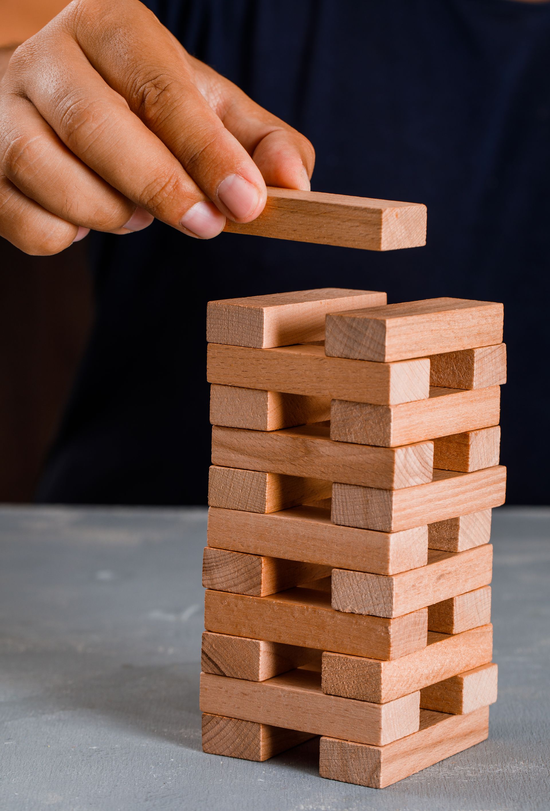 Person placing wooden block on a Jenga tower on a grey surface.