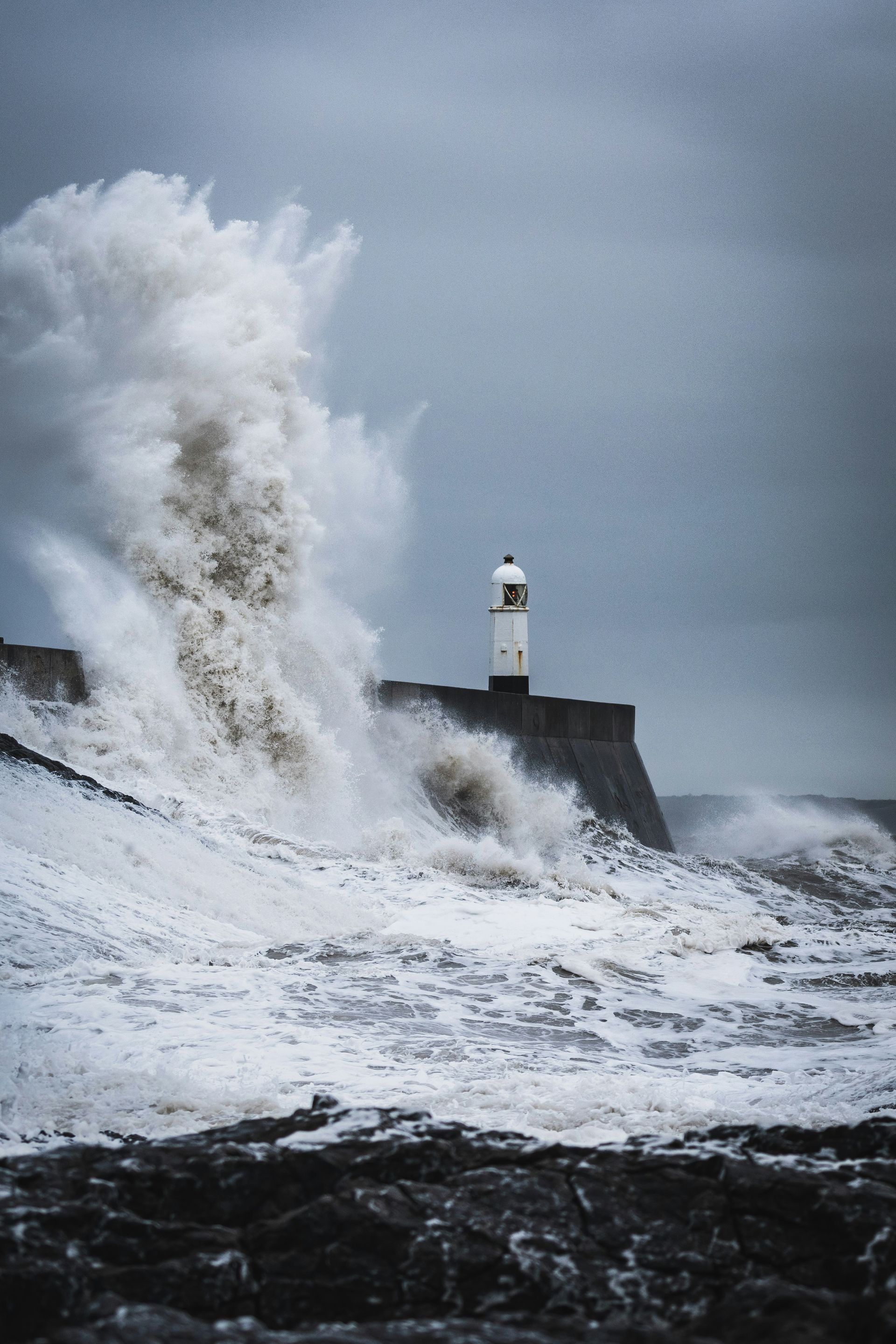 Lighthouse on a stone wall, large waves crashing during a storm under a cloudy sky.