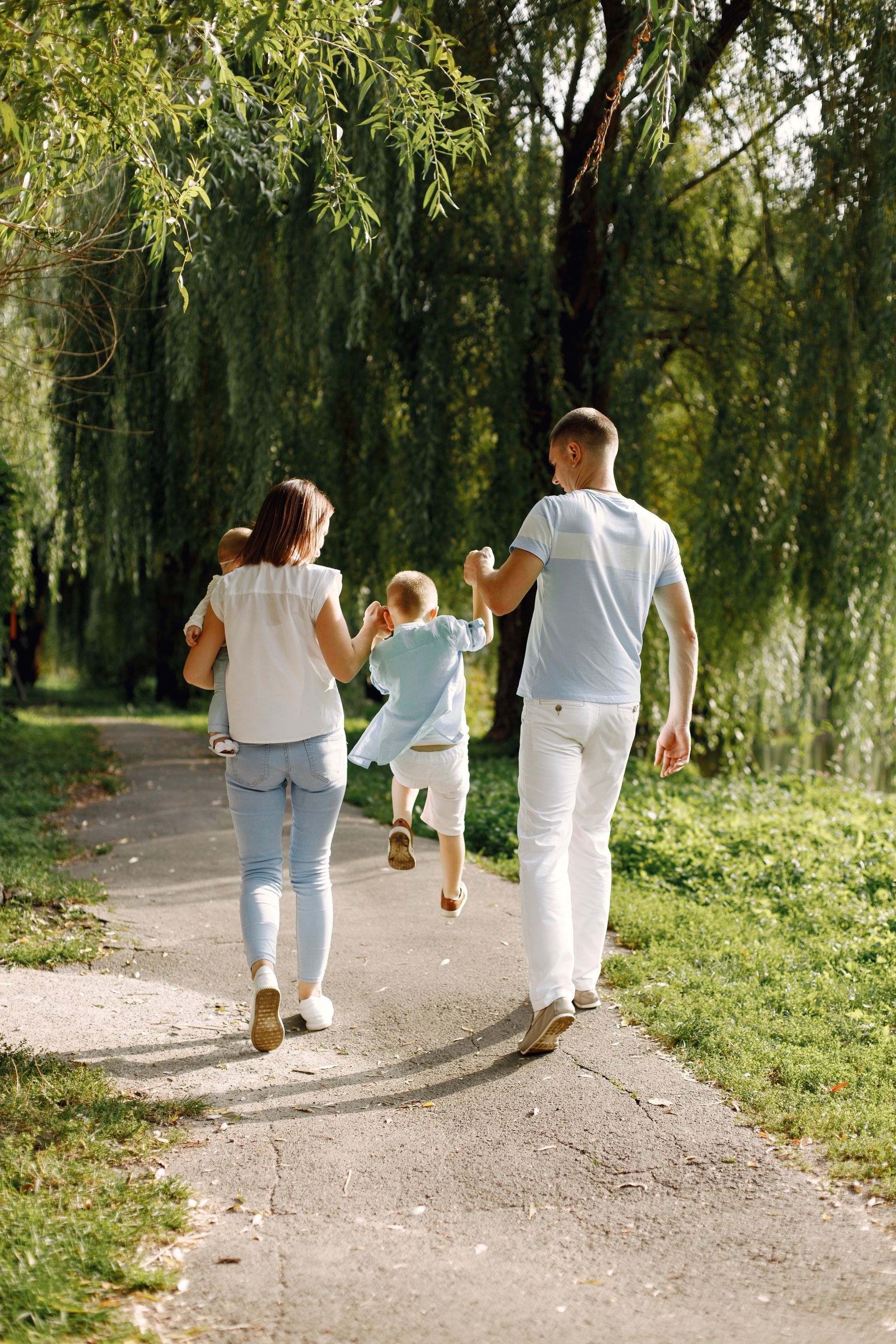 Family of four walking on a path in a park, child jumping, sunny day.